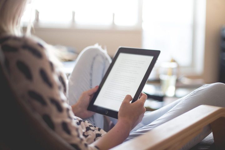 Une femme assise lit un livre numérique sur une liseuse, près d'une fenêtre