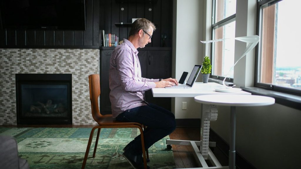 Un homme assis de profil à un bureau, les pieds au sol, le dos droit sur sa chaise, devant son ordinateur portable