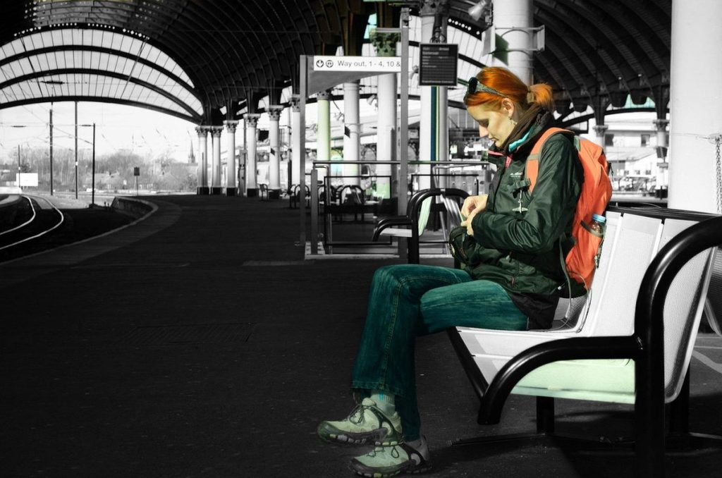 Une voyageuse avec un sac à dos orange assise sur un banc du quai d'une grande gare, sous la halle