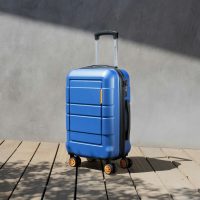 Une valise cabine rigide bleue à roulettes posée sur une terrasse en bois, devant un mur gris